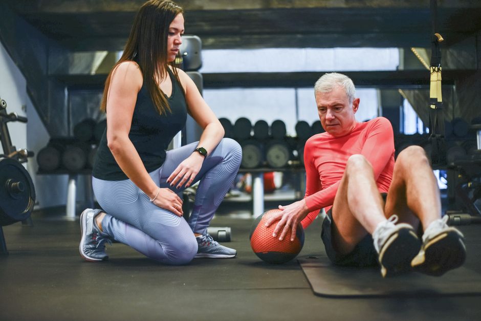 Pessoa de 60+ fazendo wall plank shoulder tap, com roupa esportiva, encostada na parede, segurando garrafa de água