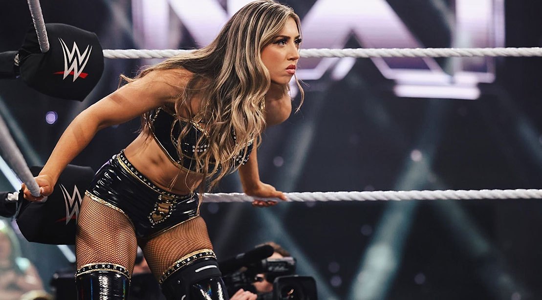 Uma mulher atlética com músculos definidos, segurando um copo de proteína e realizando um movimento de flexão de braços, enquanto está no ringue de wrestling