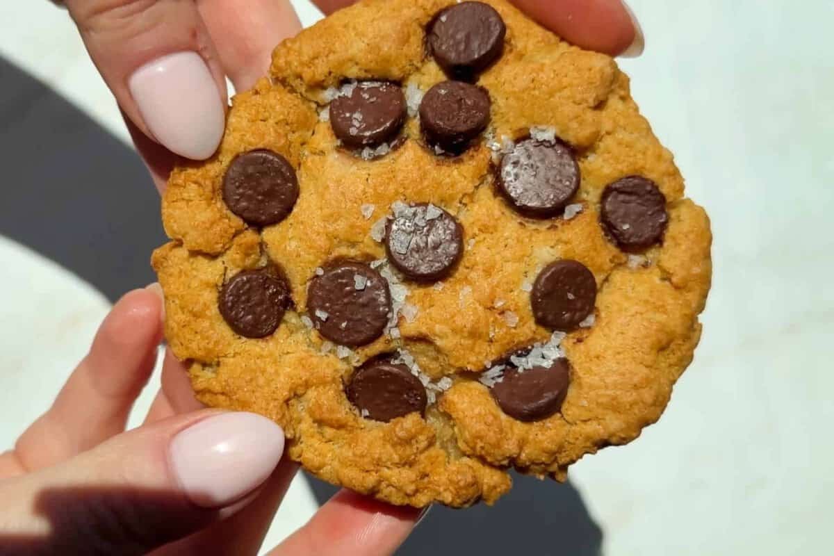 Cookie dourado saindo de uma air fryer aberta, ao lado de ingredientes frescos e uma colher de medida sobre a mesa