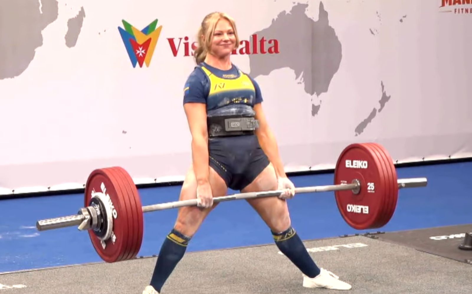 Alba Bostrom, vestindo regata esportiva, ergue uma barra carregada com discos pretos de 20 kg, cercada por piso de borracha e giz nas mãos