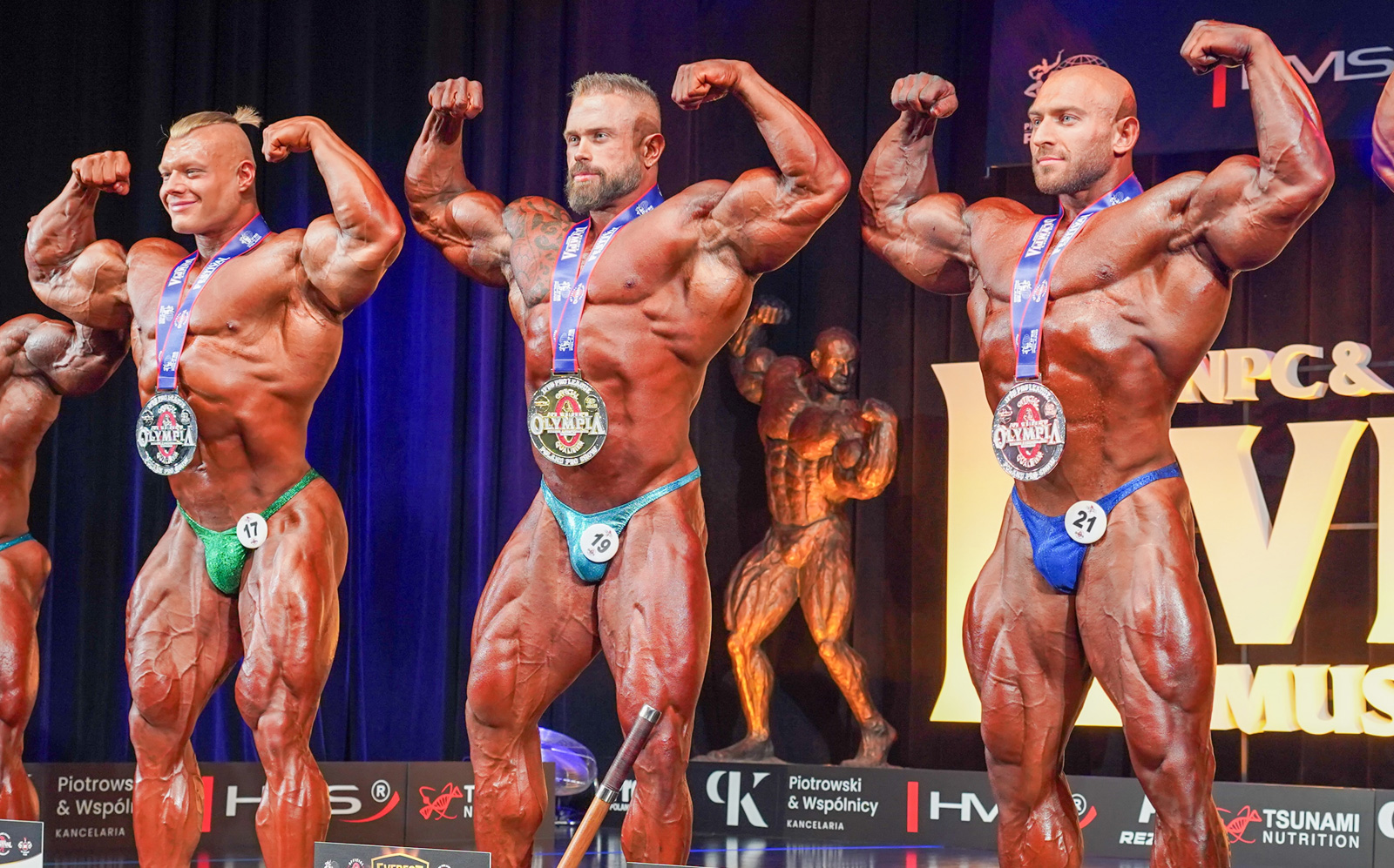 Andrea Presti musculoso, em pose de costas, segurando troféu dourado sob luzes neon e banner “Everest Muscle Legends 2026”