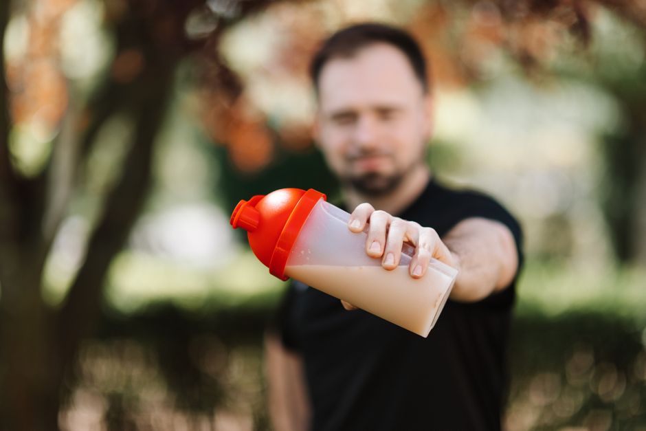 Banana, copos de whey, caseína e proteína vegetal ao lado de um liquidificador e um shake pronto em copo transparente