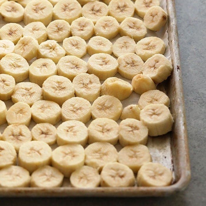 Bananas fatiadas em bandeja de silicone, prontas para ir ao freezer, ao lado de um liquidificador e folhas verdes