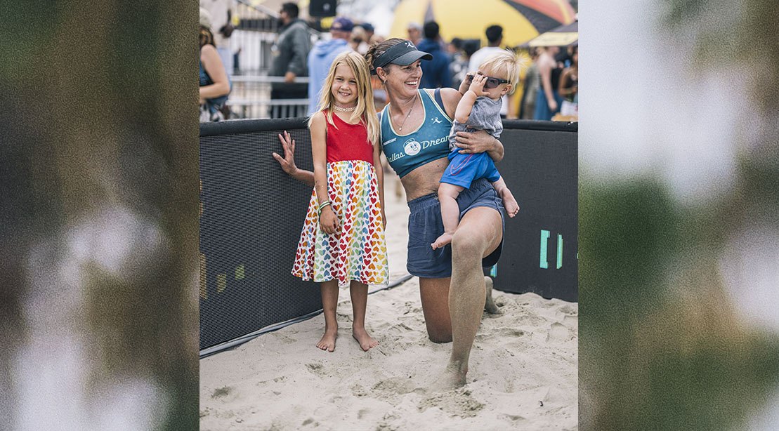 Uma mulher magra e musculada, com um bebê no colo, segurando uma bola de vôlei e sorrindo