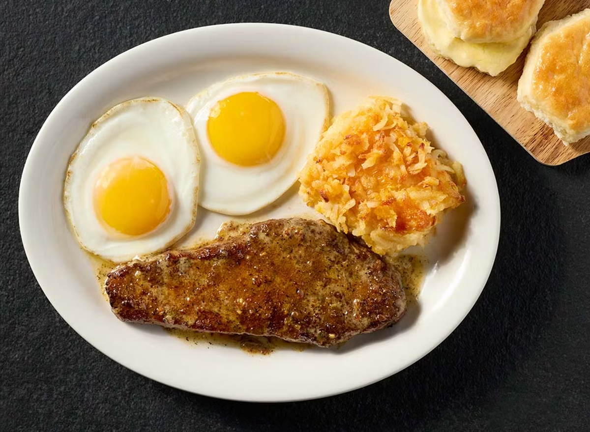 Prato com bife grelhado, ovos estalados, salada verde e vegetais coloridos sobre uma mesa de madeira bem iluminada
