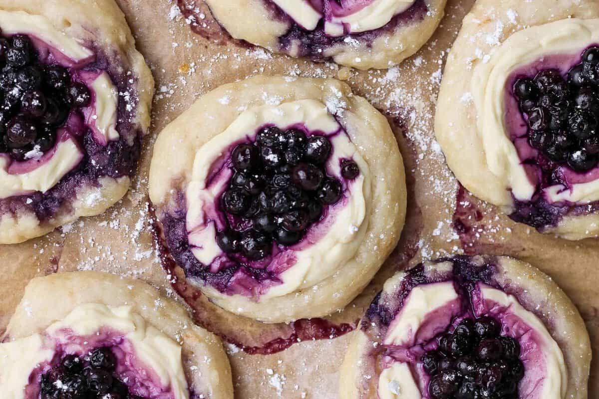 Danish de cottage cheese com blueberries frescas sobre prato branco, ao lado de copo de iogurte e colher