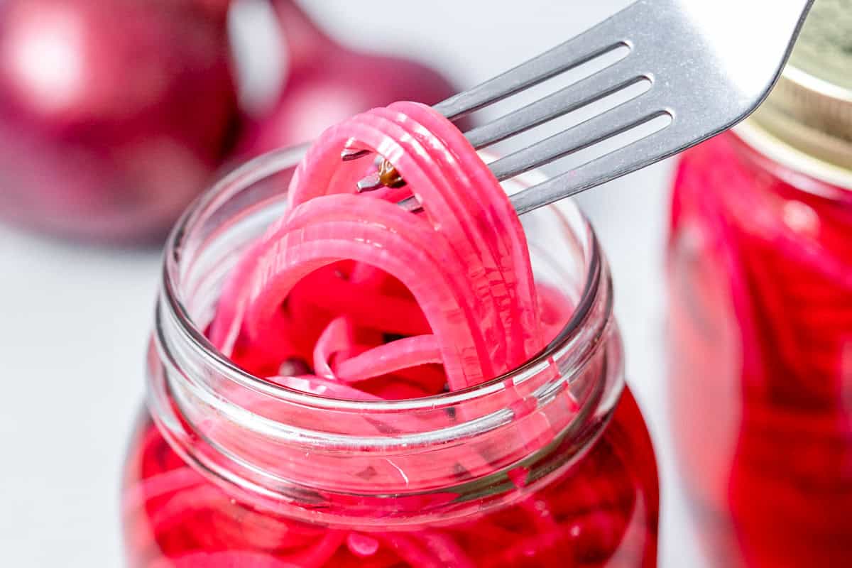 Pote de vidro com cebola roxa em conserva ao lado de uma salada fresca e um peito de frango grelhado em uma mesa
