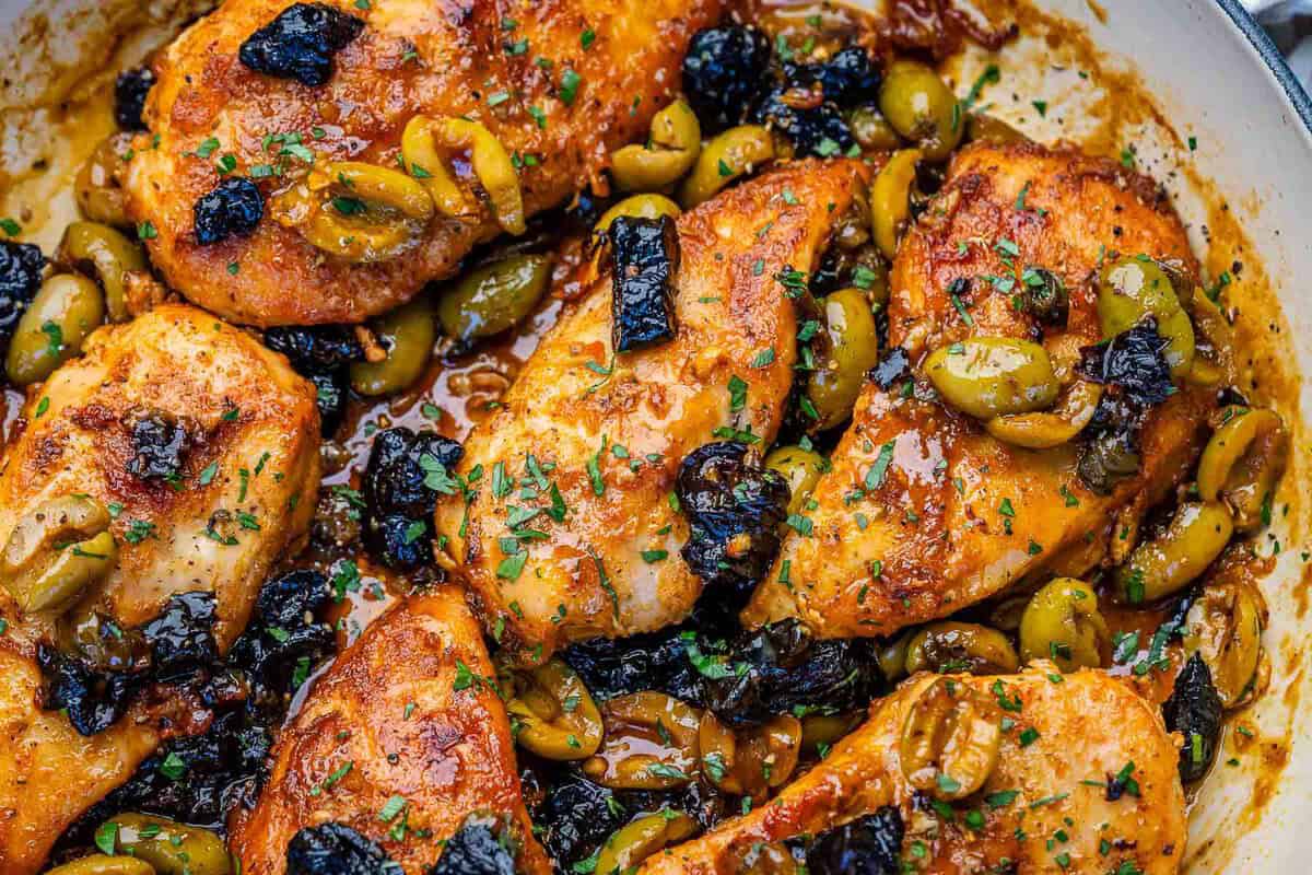 Peito de frango dourado na panela ao lado de azeitonas verdes, ameixas secas e folhas de alecrim