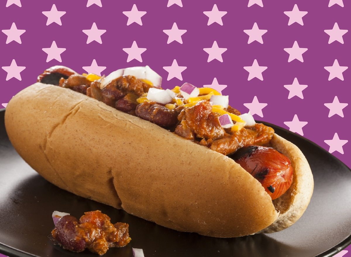 Um hot dog com molho de chili vermelho servido em uma tigela de papel com cebola e pimenta