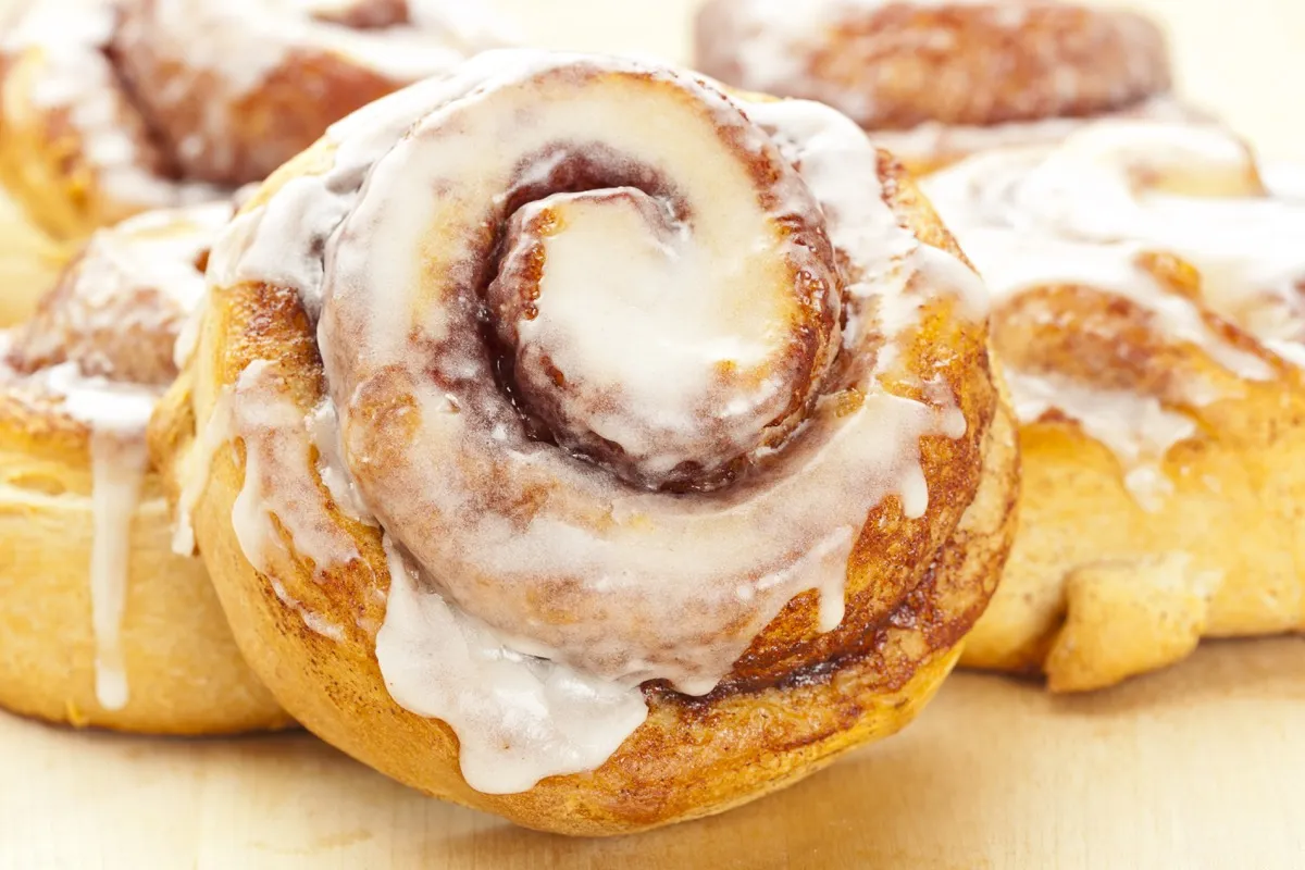 Close em um cinnamon roll dourado com calda branca, sobre uma mesa de madeira com canelas em pau e um café ao lado