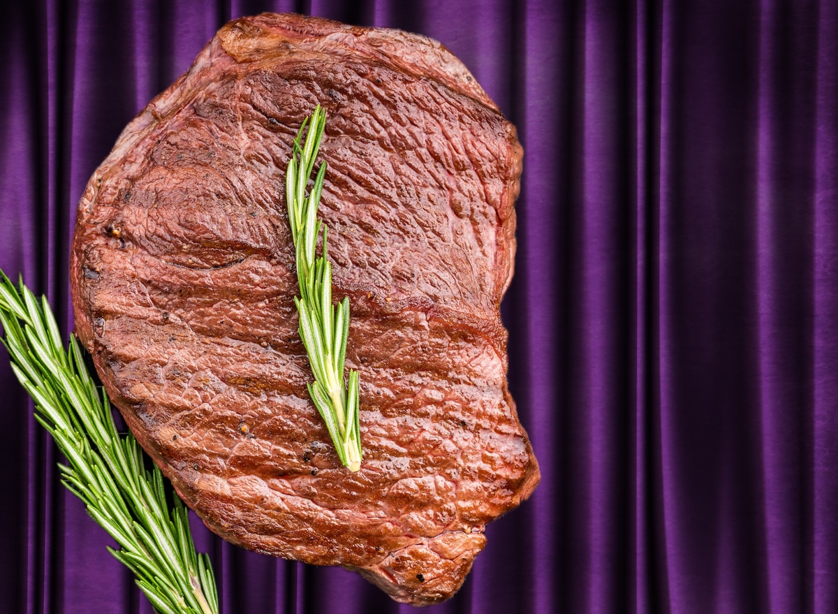 Corte de carne bovina premium cru ao lado de uma batata assada recheada e ervas frescas sobre uma bancada de madeira