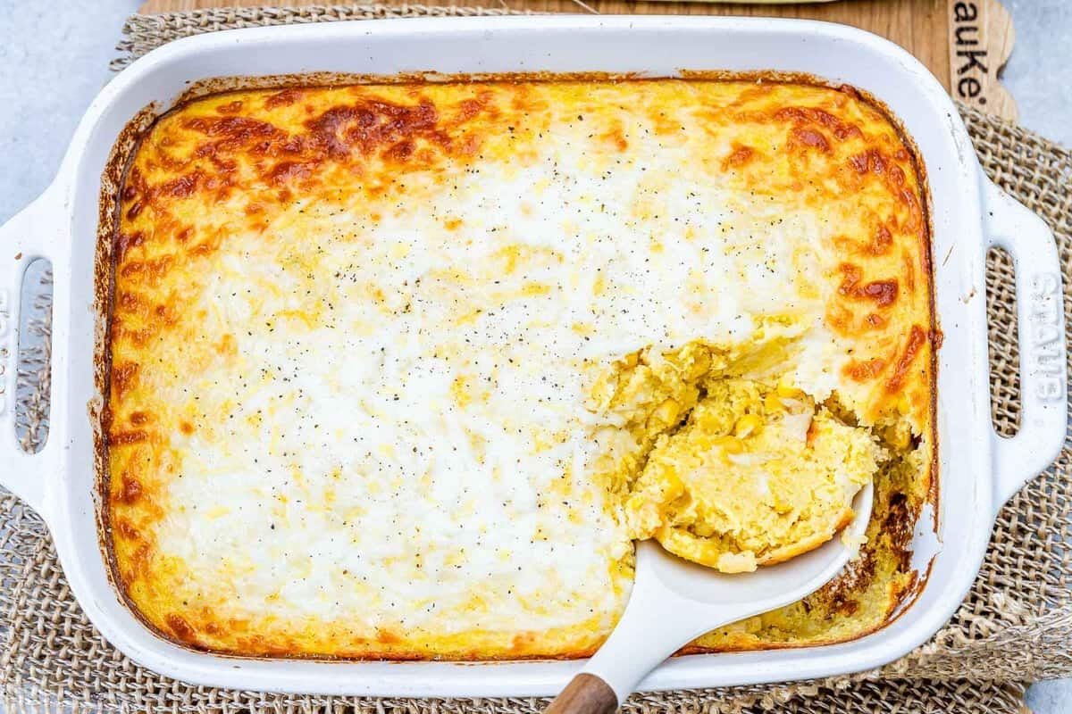 Gratinado dourado de milho cremoso com iogurte, queijo derretido e ervas, servido em travessa de cerâmica ao lado de uma colher