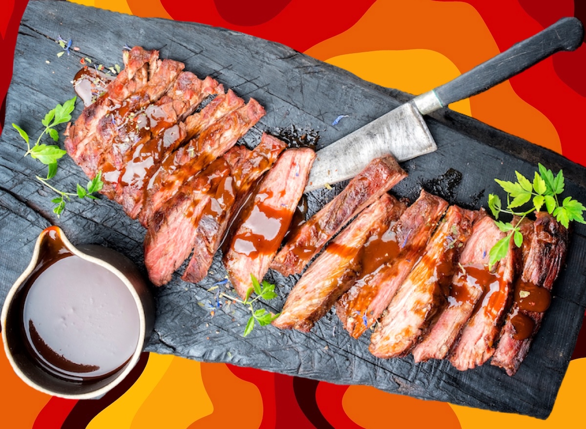 Um pitmaster segurando um corte de carne marmorizado, com um pedaço de flank steak e um churrasqueiro ao fundo