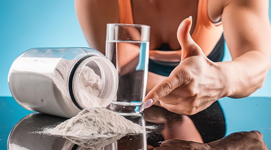 Mulher musculosa segurando halteres, ao lado de pote de creatina e copo de água, em academia bem iluminada