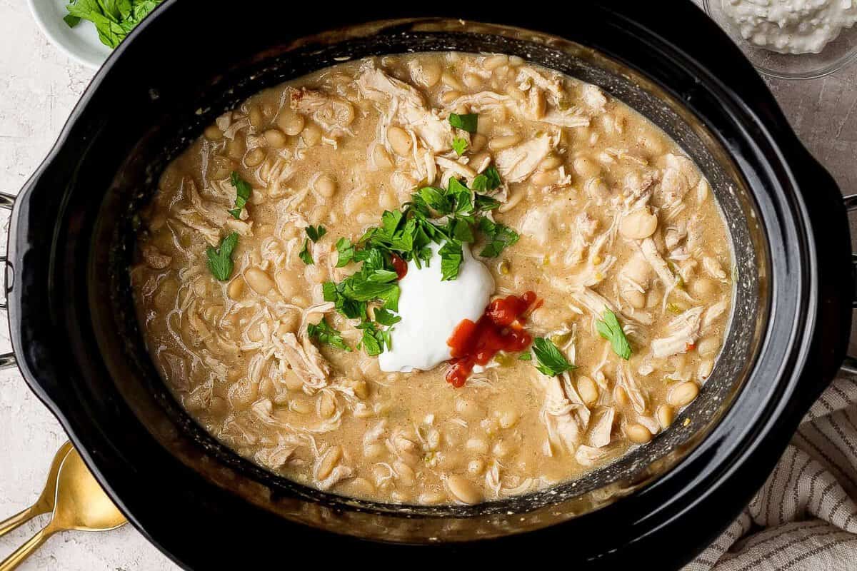 Frango desfiado, feijão manteiga e milho amarelo fervendo em uma crockpot branca, cercados por pimentões verdes e limão