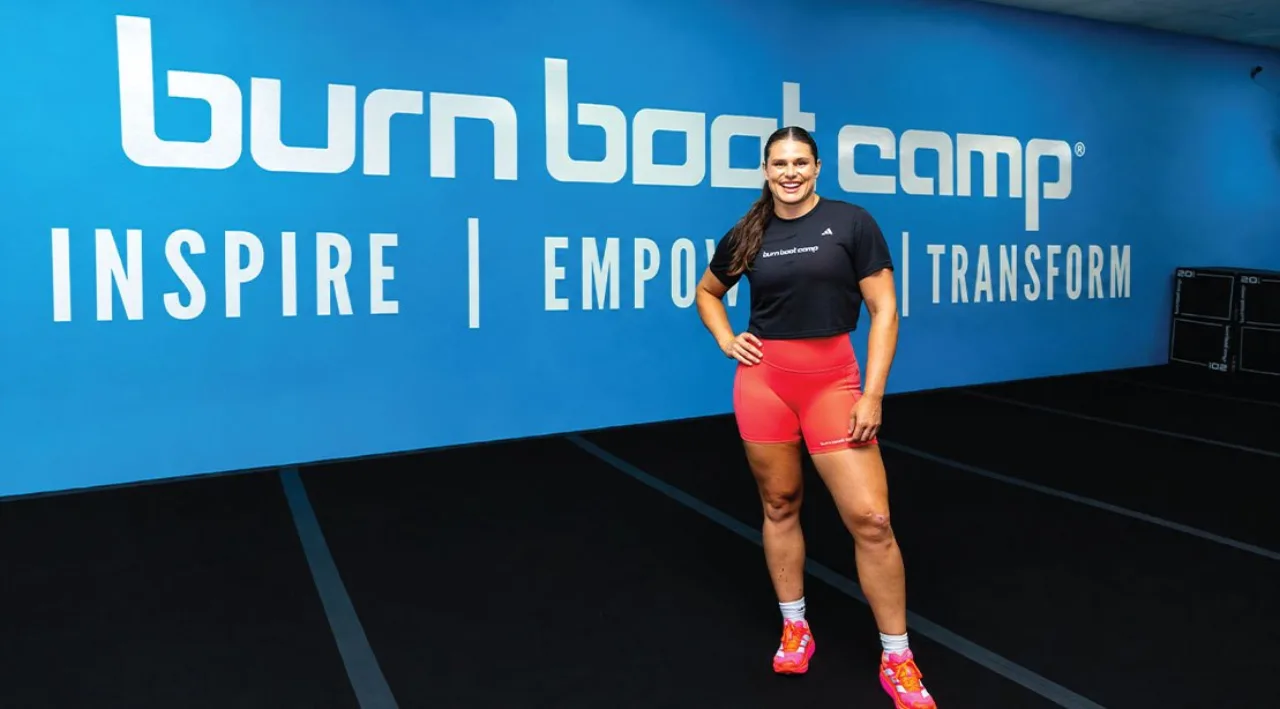 Ilona Maher vestindo uniforme de rugby, segurando bola, ao lado de halteres e cronômetro, em pista de atletismo