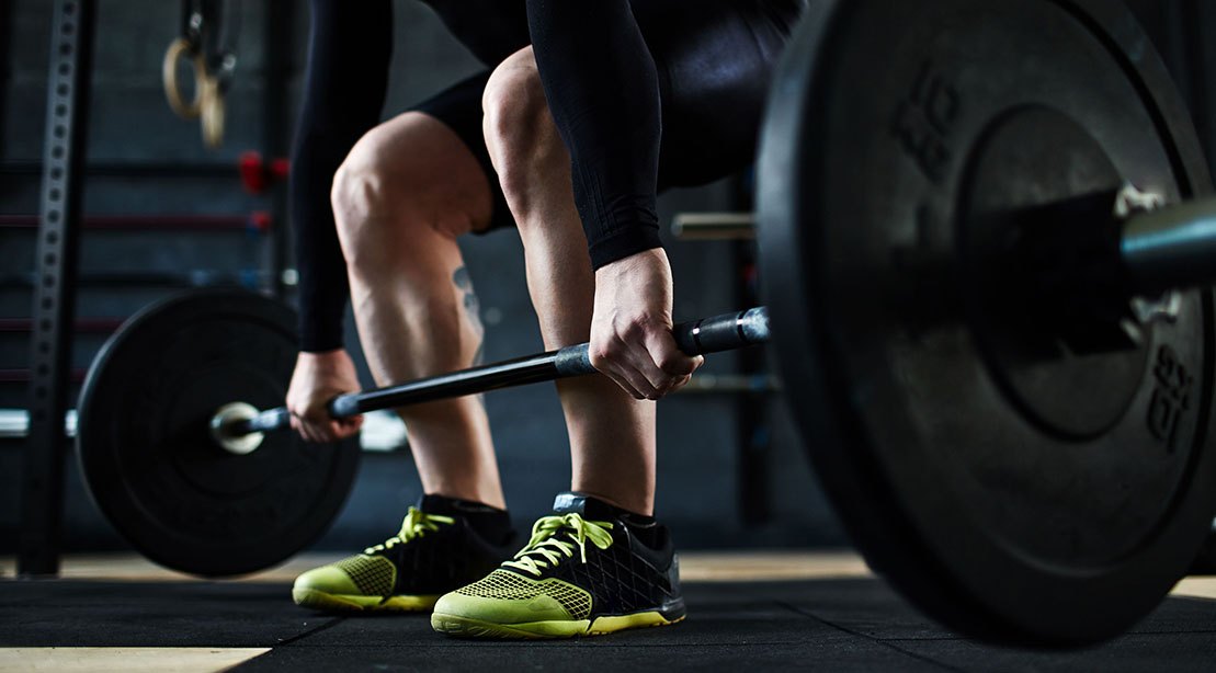 Atleta em roupa de ginástica faz deadlift com barra, costas retas, quadris alinhados e joelhos levemente dobrados