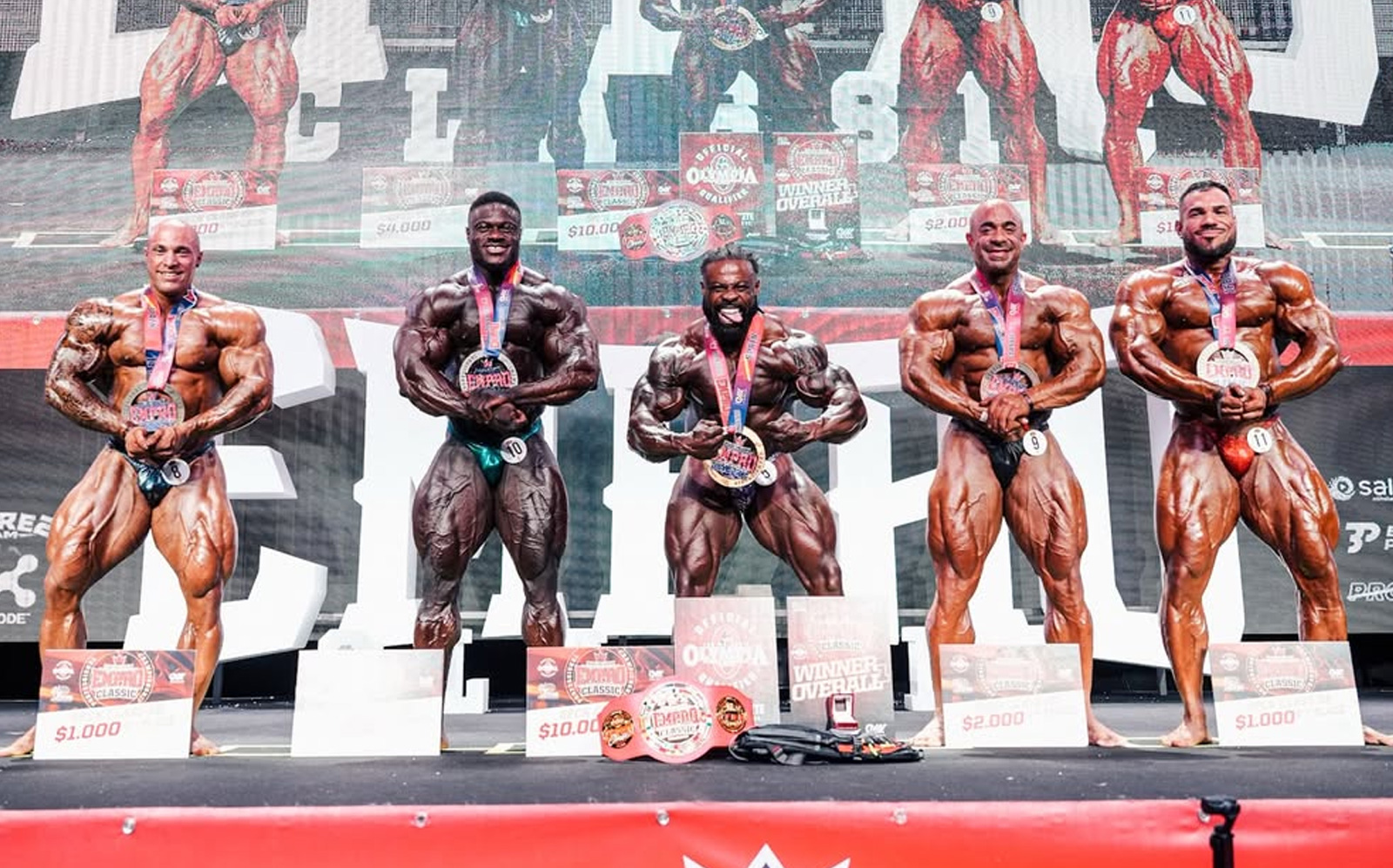 Atletas musculosos posam no palco iluminado, segurando troféus enquanto bandeiras da Espanha e da IFBB ao fundo
