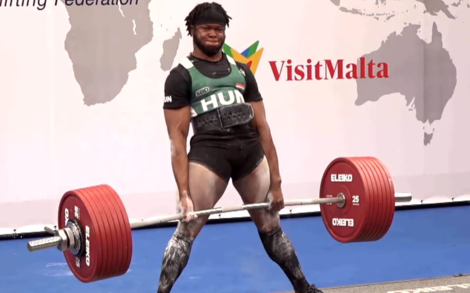 Enahoro Asein executa deadlift com barra carregada de discos de aço, usando luvas de couro e chão de borracha