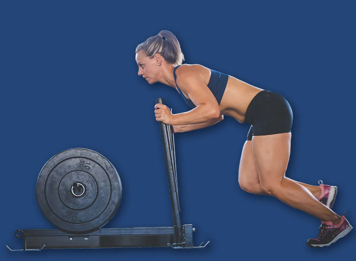 Mulher em pé, realizando um exercício de levantamento de peso com um bastão ou barra, com legumes frescos ao fundo