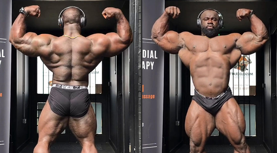 Samson Dauda posando em um palco de fisiculturismo, exibindo volume muscular extremo e definição sob luzes intensas