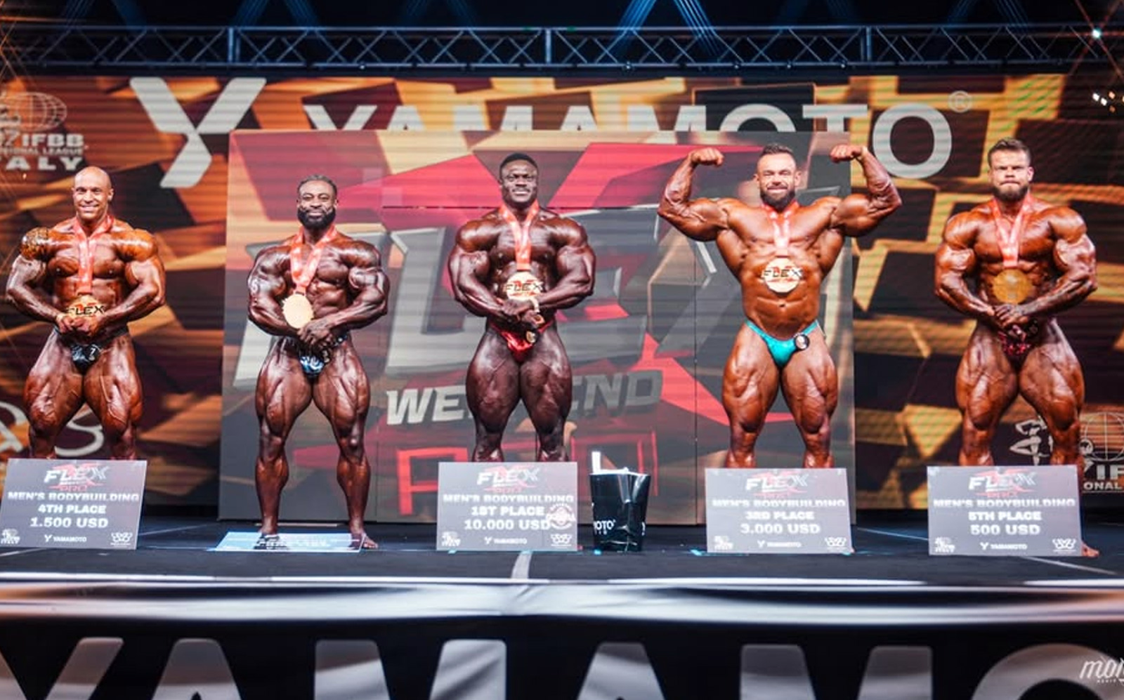 Um homem musculoso em primeiro lugar, segurando um troféu e sorriente, ao lado de dois outros corpos magros