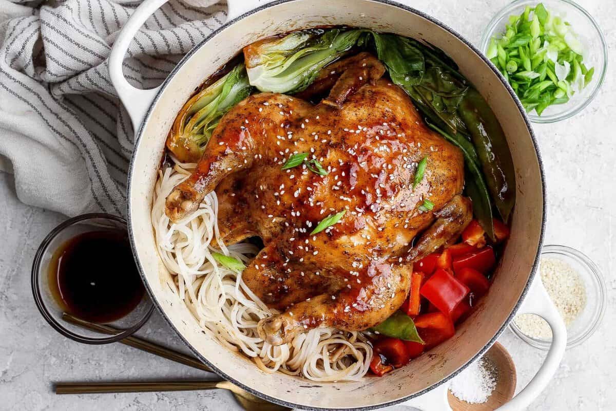 Frango inteiro dourado na panela, rodeado de legumes coloridos (pimentão, brócolis e cenoura) e molho brilhante de stir‑fry