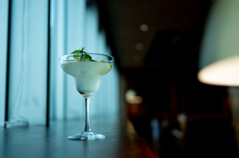 Pessoa segurando um copo de frozen margarita ao lado de um liquidificador, limões cortados, garrafa de tequila e xarope de agave