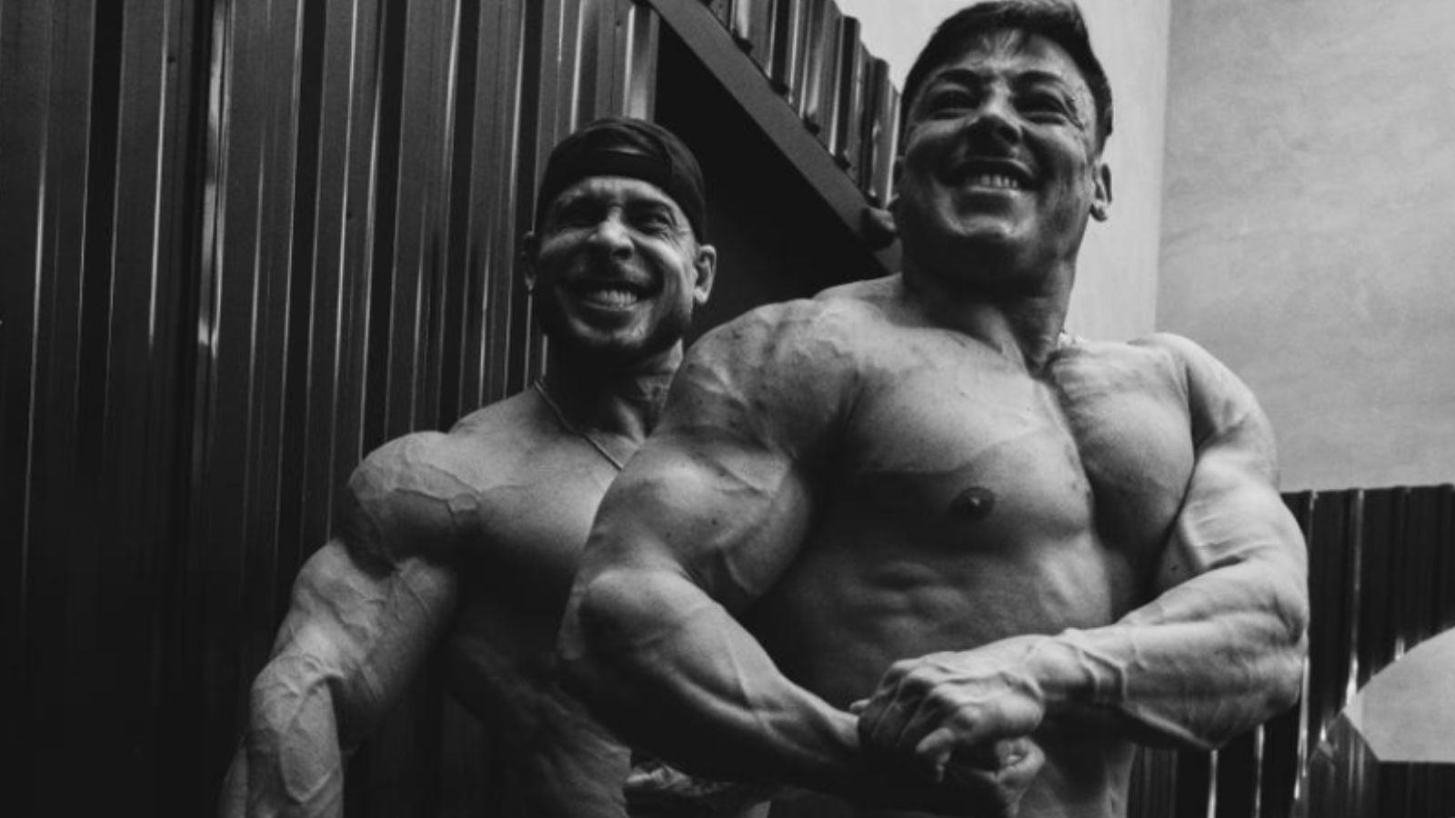 Atleta fisiculturista posando sob luz dramática em academia com halteres e espelhos ao fundo, focando na musculatura