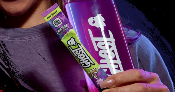 Um homem em trajes de academia, com um copo de café em uma mão e um saco de GHOST Energy Sticks na outra
