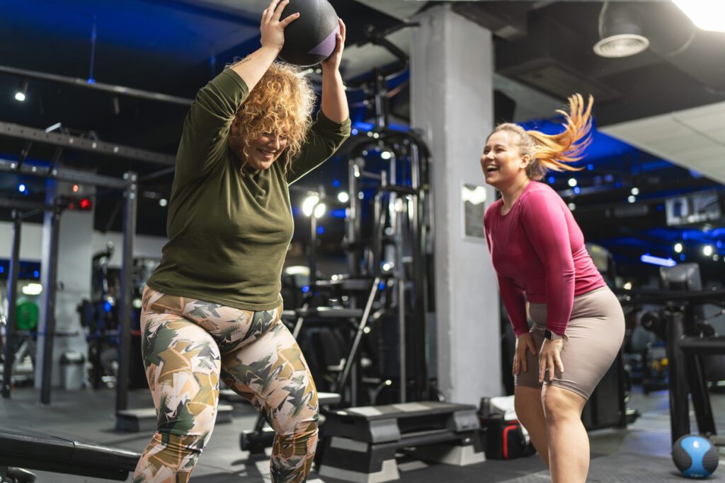 Mulher em roupa de treino segurando um shaker de proteína ao lado de halteres, olhando para um relógio de pulso