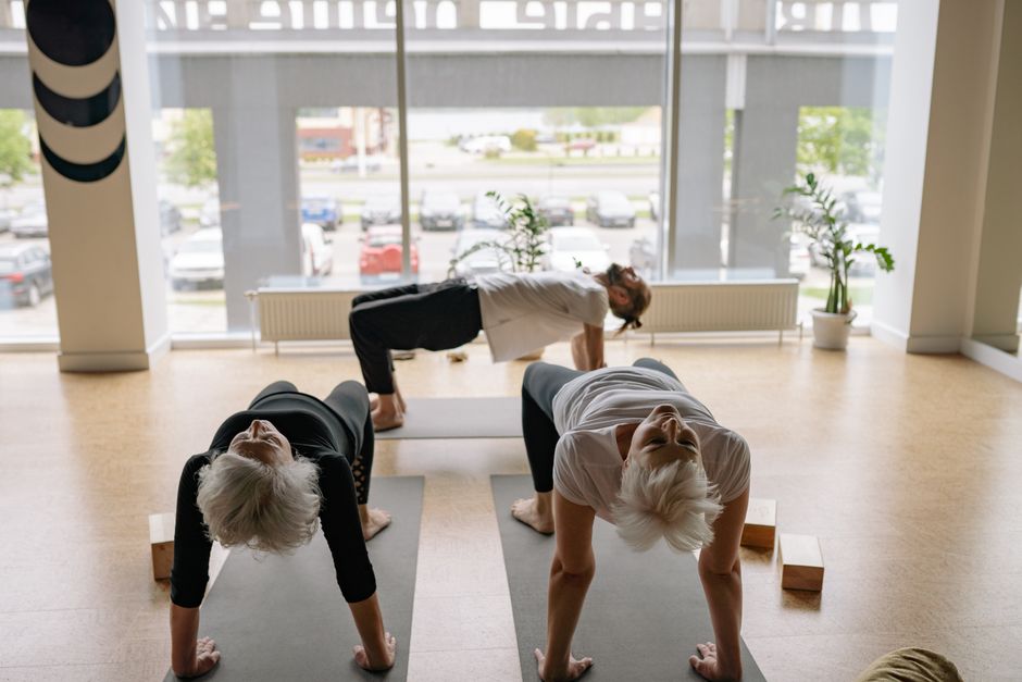 Uma pessoa fazendo glute bridge em uma academia, com pesos na cabeça e um personal trainer assistindo no fundo