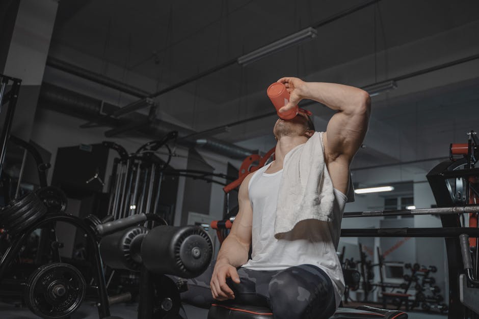 Atleta bebendo água em uma garrafa squeeze ao lado de halteres e uma toalha em uma academia bem iluminada