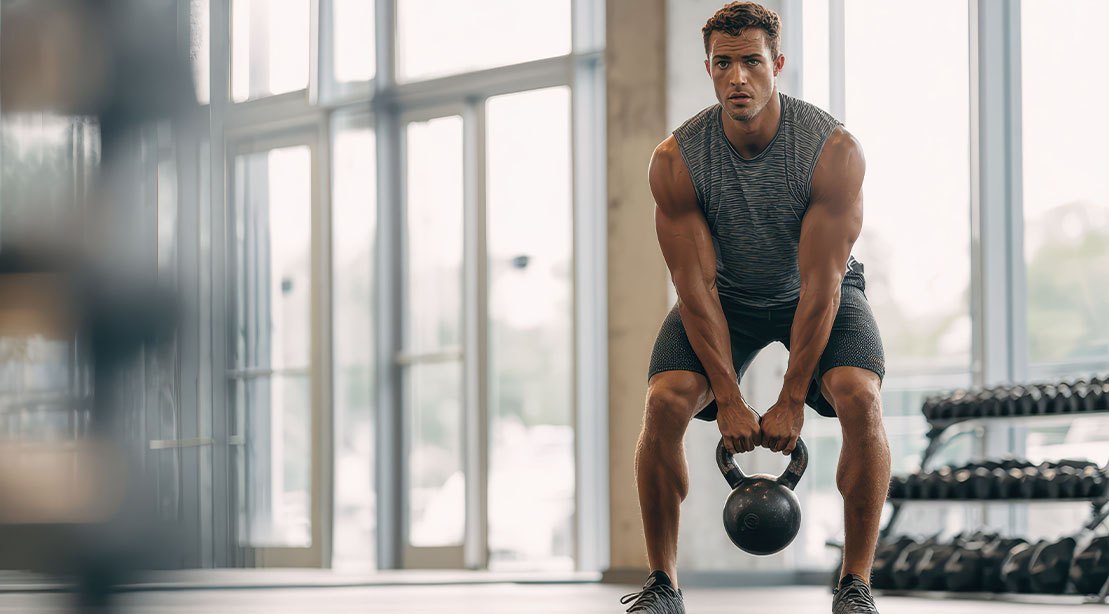 Mulher em roupa esportiva faz hip hinge ao segurar kettlebell, com parede ao fundo marcando a distância dos pés