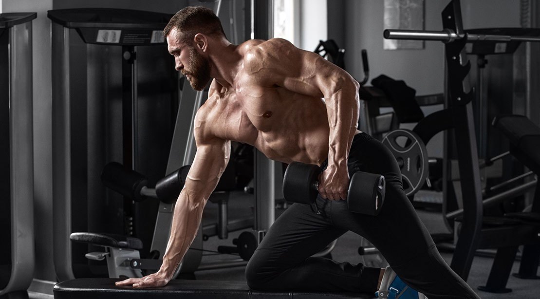 Homem musculoso segurando shaker de whey ao lado de halteres e um prato com peito de frango grelhado