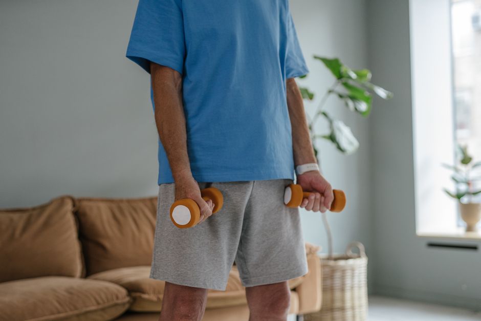 Idoso(a) em pé, usando faixa elástica nos pés e segurando as pontas para fazer curls, ao lado de um copo de shake de proteína