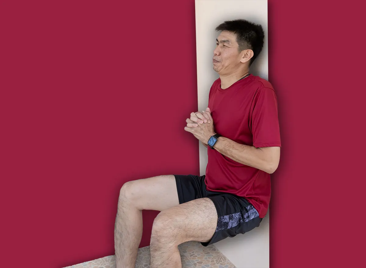 Pessoa idosa sorridente realizando o exercício de wall sit com as costas apoiadas em uma parede neutra