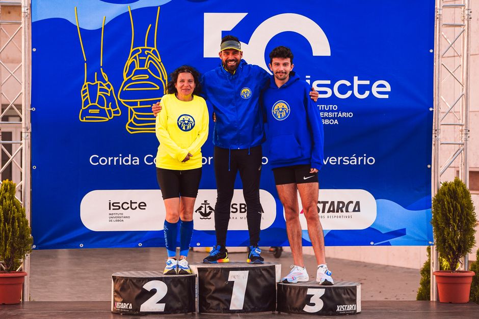 James Sprague e Emma Tall no pódio, segurando medalhas enquanto exibem músculos definidos e placas de pontuação ao fundo