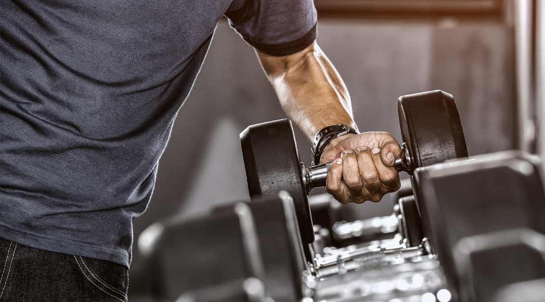 Uma pessoa segurando um kettlebell e uma dupla de dumbbells ao lado de uma parede de blocos de pesos