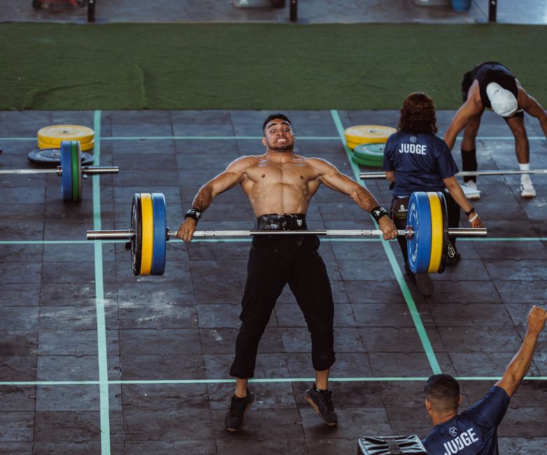 Atleta musculoso ergue um log de ferro de 210 kg sobre a cabeça, cercado por árbitros e plateia vibrante