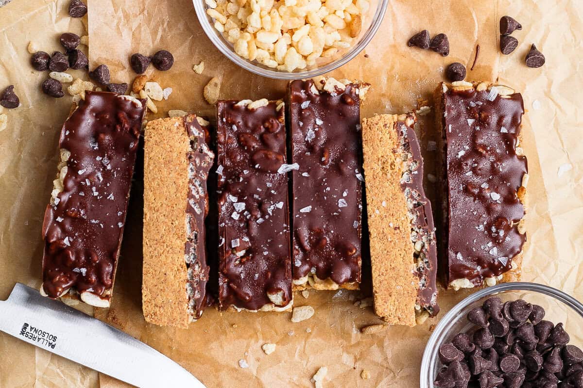 Barra de proteína sobre tábua, ao lado de manteiga de amêndoas, xarope de bordo, whey em pó e nozes, com garrafa de água