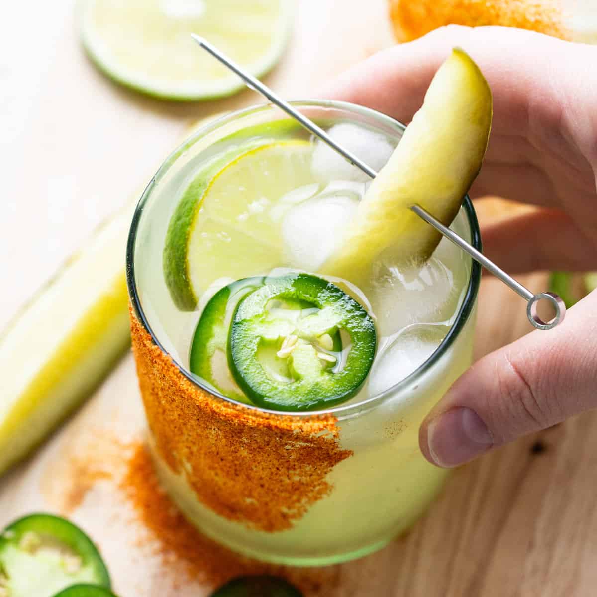 Margarita de picles com borda de sal e pimenta ao lado de um pepino fresco sobre uma mesa de madeira rústica