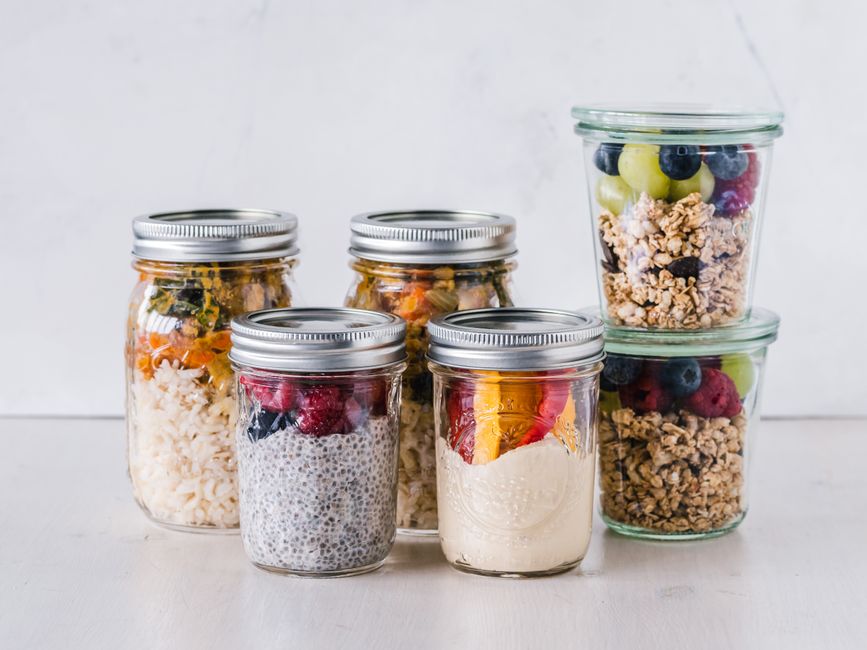 Uma mulher preparando uma salada de frutas e legumes em um recipiente de plástico transparente, ao lado de uma lista de ingredientes e um relógio de cozinha