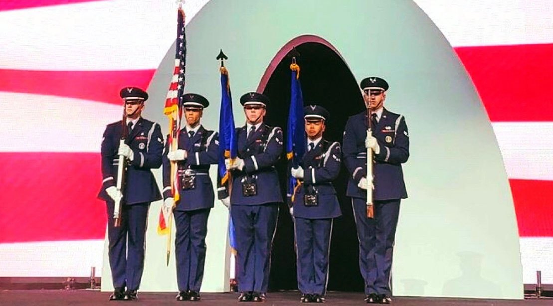 Atleta musculoso em pose de competição usando farda militar, segurando halteres e ao fundo bandeira das forças armadas