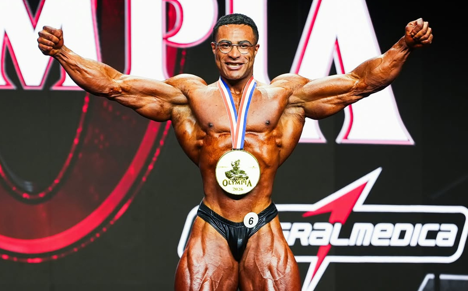Niall Darwen em pose de Classic Physique, segurando o troféu da Olympia, ao lado de halteres e bandeira britânica
