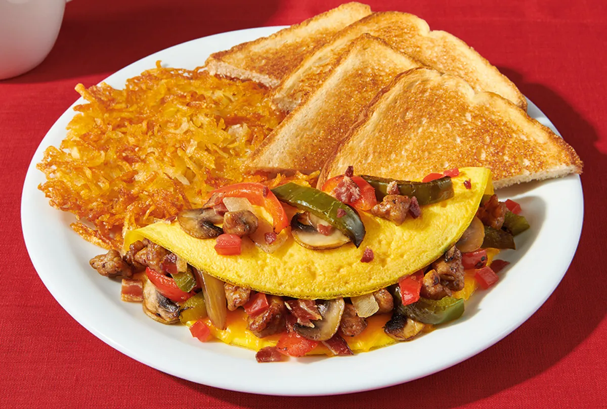 Omelete dourada recheada com espinafre e tomate ao lado de talheres em uma mesa de madeira bem iluminada
