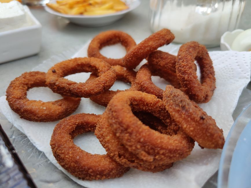 Cebola em rodelas empanadas ao lado de uma air‑fryer, com folhas de rúcula e um copo de água gelada
