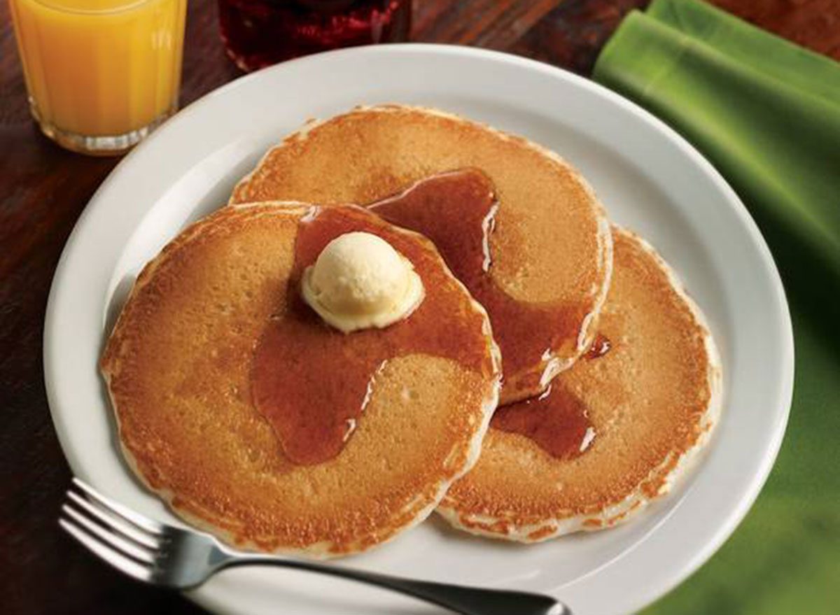 Mulher em roupa de ginástica segurando pilha de pancakes com mel, morangos e manteiga ao lado de um copo de água