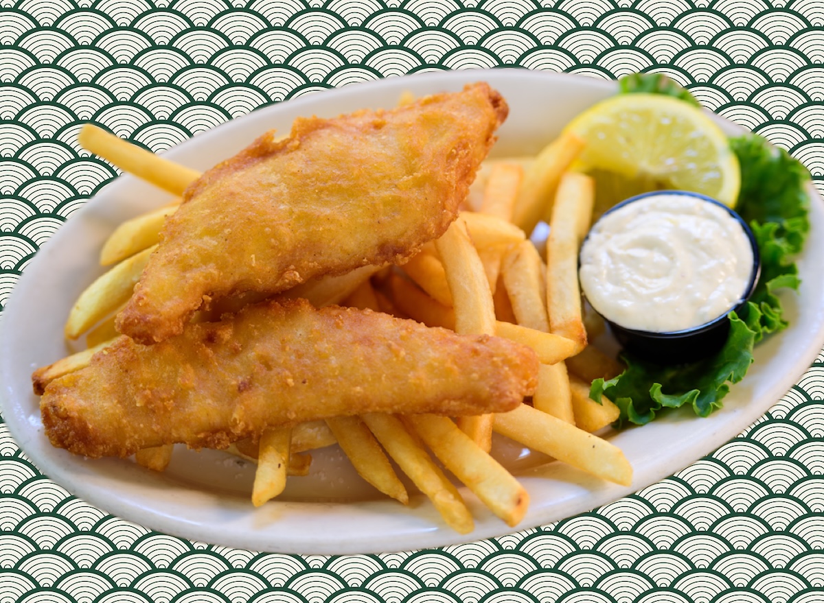 Filé de peixe dourado e crocante sobre uma tábua de madeira, acompanhado de limão fatiado e ervas frescas