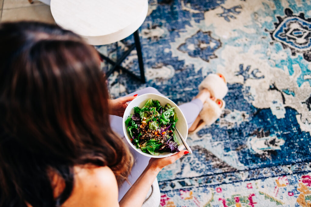 Mãos organizando marmitas com vegetais frescos, grãos e frutas sobre uma mesa de cozinha com lista de compras ao lado